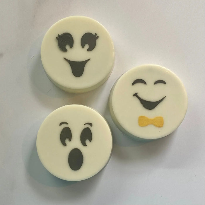 A white marble countertop with three white chocolate covered Oreos decorated with the three faces on the Ghost Faces Cookie Stencil in black royal icing with the bow tie feature made in orange, BakeArt Stencils