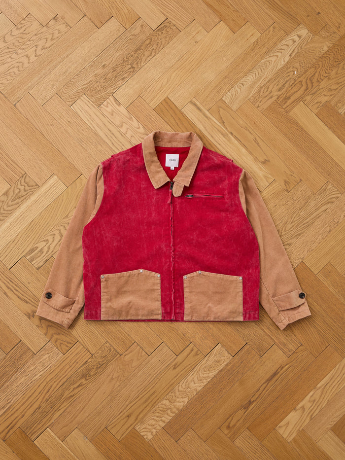 The Found Corduroy Field Jacket features a two-tone design with tan sleeves and collar, a red corduroy front, tan pockets, zippered chest pocket, and buttoned cuffs, shown laid flat on a wooden herringbone floor.