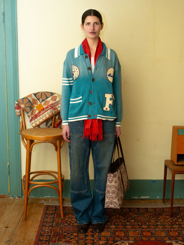 A person in a room wears the Denim Carpenter Jeans by FOUND—relaxed straight-leg and patched blue cardigan, accessorized with a red scarf, holding a patterned tote. Beside them, a wooden chair draped with a quilt sits on a patterned rug.