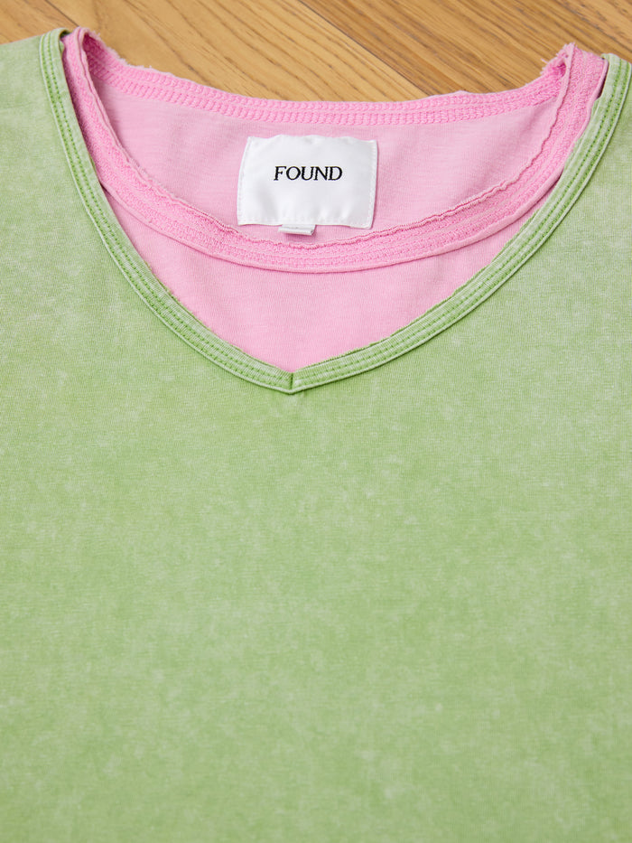 A close-up of the FOUND Double Layer LS Tee in green sunfade wash V-neck, layered over a pink crew neck with a white "FOUND" label, displayed on a wooden surface.