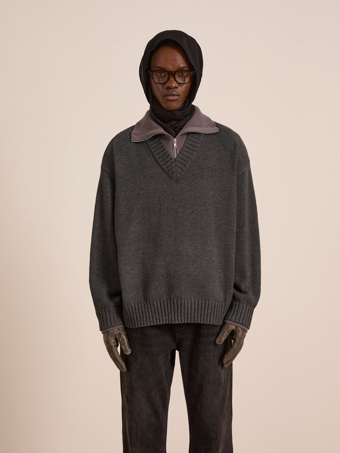 A person in FOUND’s Double Layer Sweater—an oversized gray wool blend V-neck layered over a collared shirt—paired with dark jeans, gloves, a black headscarf, and dark glasses stands against a plain light background.
