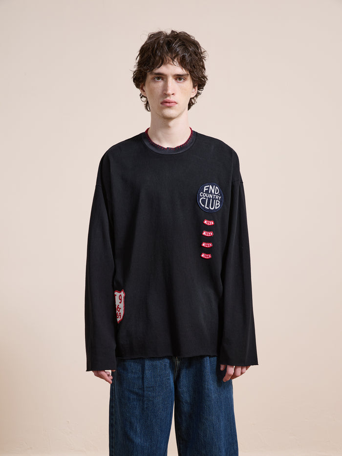 A person with medium-length curly hair wears the FOUND Embroidered Patches LS Tee, a vintage washed black unisex relaxed fit long-sleeve shirt, with blue jeans, standing against a plain beige background.