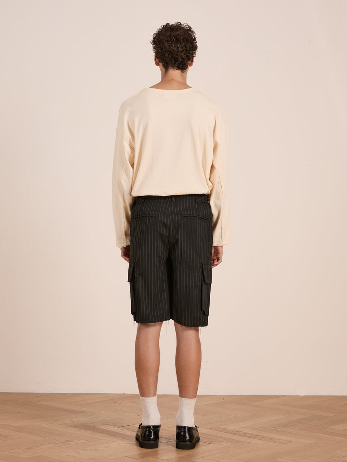 A person stands indoors facing away, wearing a cream long-sleeve shirt, FOUND Hava Pinstripe Pleated Cargo Trouser Shorts with an adjustable fit, white socks, and black shoes.