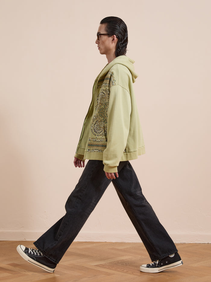 Wearing a light green FOUND Motif Zip-Up Hoodie, black flared jeans, and black sneakers, someone walks across a wooden floor by a beige wall—showcasing iconic streetwear style.
