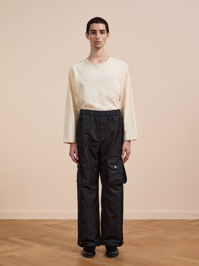 A person stands indoors on a wooden floor, wearing FOUND's black Nylon Cargo Pants—unisex with side pockets and a relaxed fit—paired with a cream long-sleeve shirt, facing the camera against a beige wall.