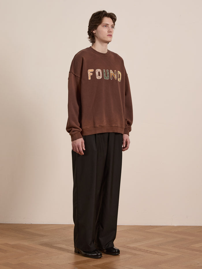 A medium-haired person stands against a plain backdrop, wearing a FOUND Patterned Logo Crewneck made from French terry cotton with "FOUND" appliqué lettering and loose dark trousers, looking to the side.