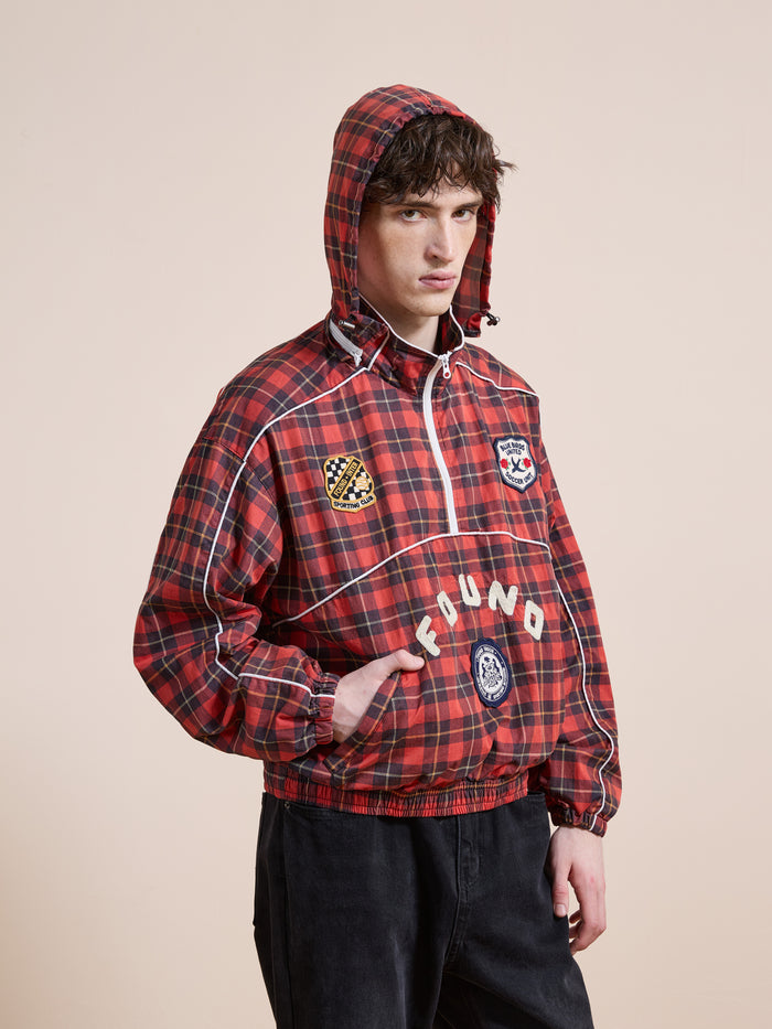Someone stands against a plain beige background wearing the FOUND Red Plaid Track Jacket, a unisex relaxed-fit piece featuring multiple patches and the brand name "FOUND" on the front.