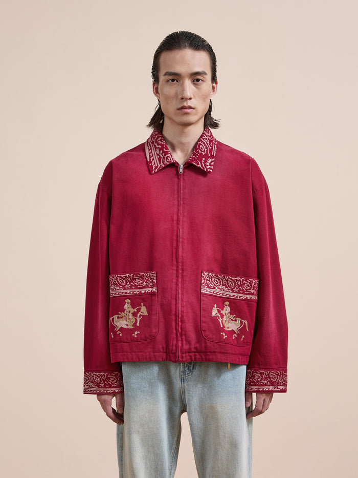 A person wears the Found Abhala Printed Work Jacket, featuring embroidered equestrian motifs and a unisex relaxed fit, paired with light blue jeans against a beige background.