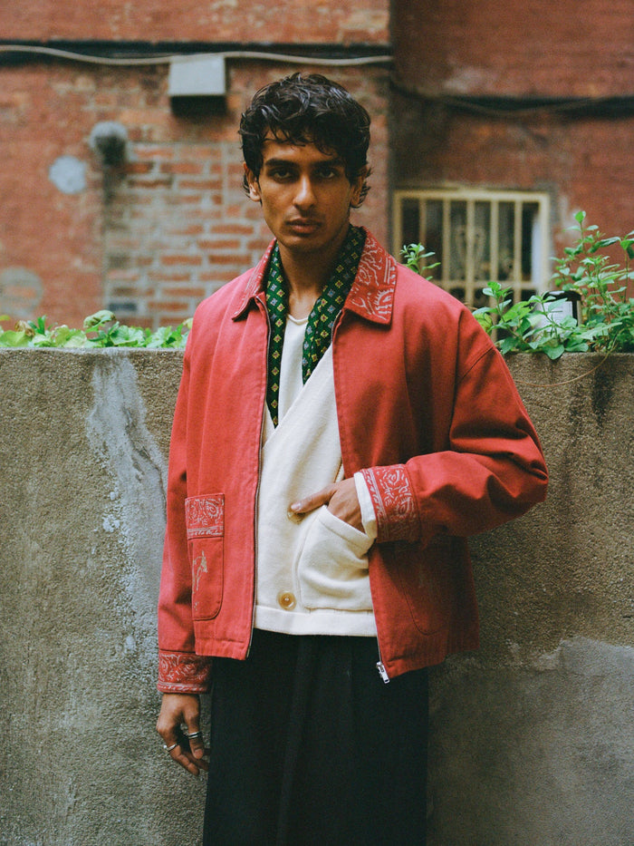 A person stands outdoors by a concrete wall, wearing Found’s Abhala Printed Work Jacket with equestrian motifs, layered over a white shirt and black pants. The unisex jacket offers a relaxed fit, and the individual has a neutral expression.