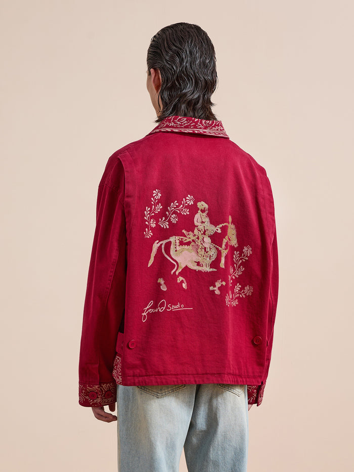 A person faces away, wearing Found’s Abhala Printed Work Jacket with a unisex relaxed fit, floral embroidery, and equestrian motifs on the back, paired with light blue jeans against a plain background.