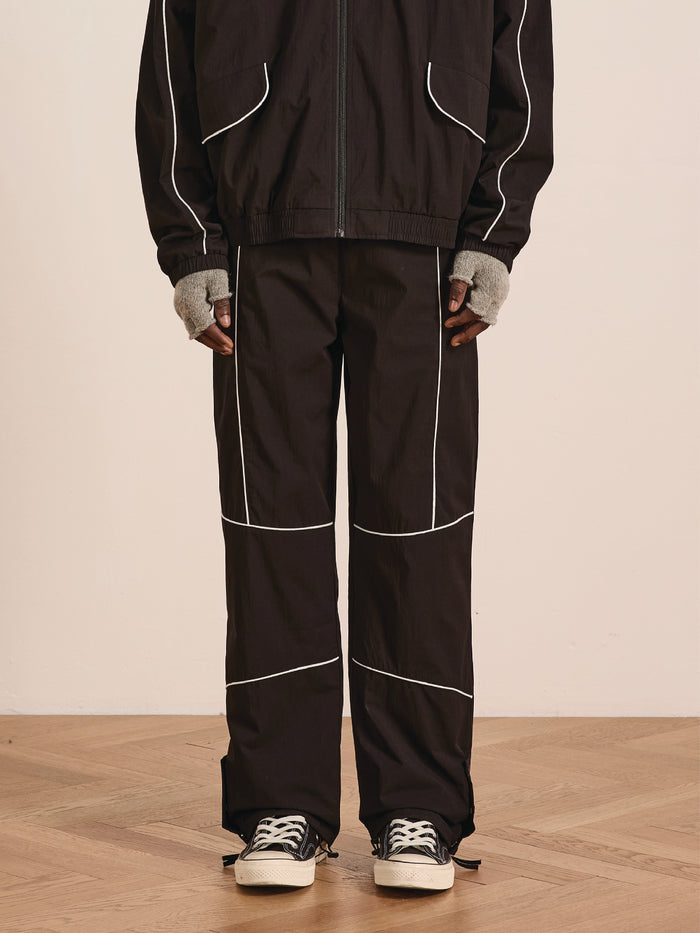 A person stands on a wooden floor wearing Found's Arena Stripe Track Pants—black with white piping and an adjustable waistband—paired with a black zip-up jacket, gray fingerless gloves, and black sneakers for a unisex sportswear look.
