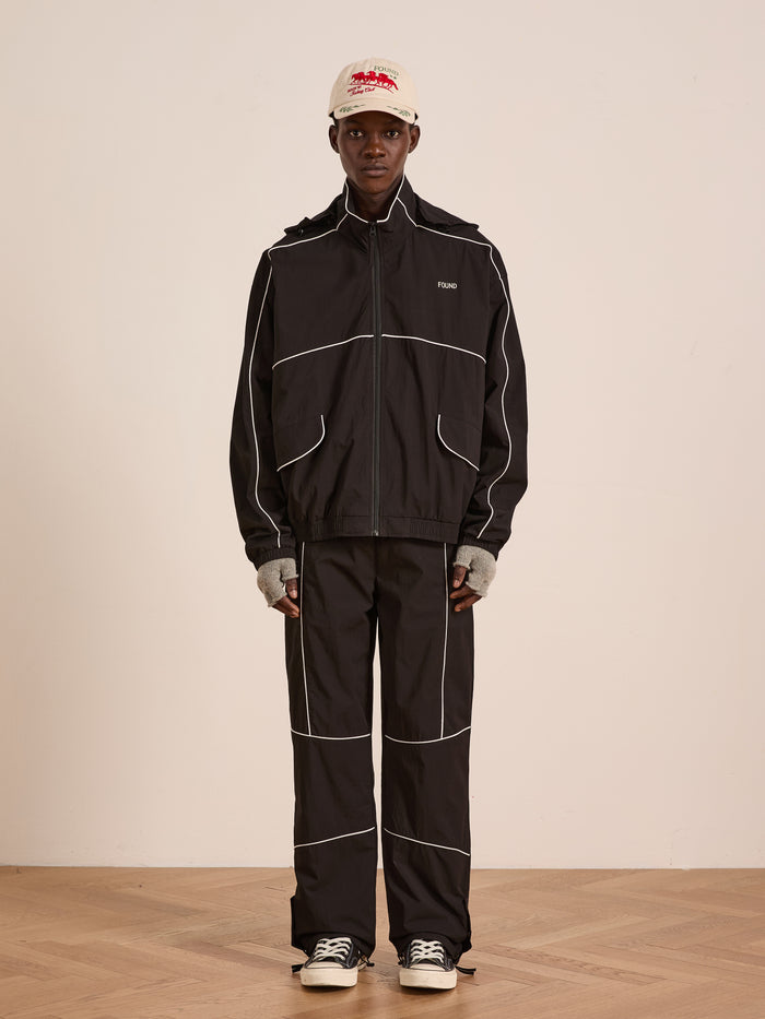 A person stands indoors in Found’s Arena Stripe Track Pants—a black pair with white piping and an adjustable waistband—styled with a beige cap, gray fingerless gloves, and black-and-white sneakers against a plain background.