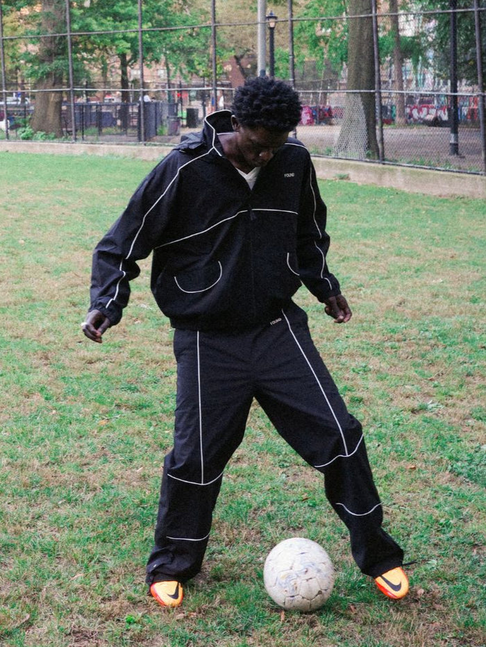Wearing Found’s Arena Stripe Track Pants, a person stands on a grassy field in relaxed style, looking down as they control a soccer ball with their foot—highlighting fashionable unisex sportswear.