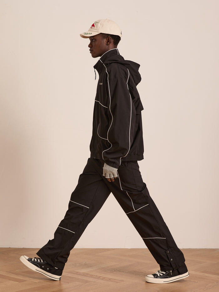 A person walks indoors in a black jacket, white cap, gray gloves, and black Converse sneakers. Their look features Found's Arena Stripe Track Pants—unisex sportswear—standing out against a simple, neutral background.