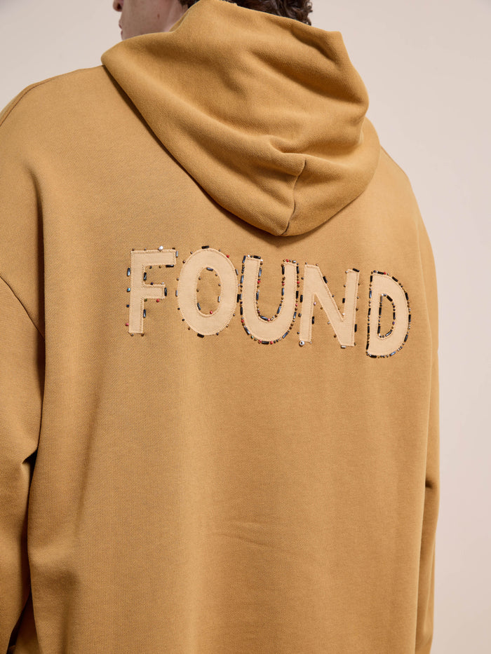Model wears the FOUND Beadwork Logo Hoodie, a unisex relaxed-fit style in tan french terry with "FOUND" stitched large on the back, finished with a vintage enzyme wash.
