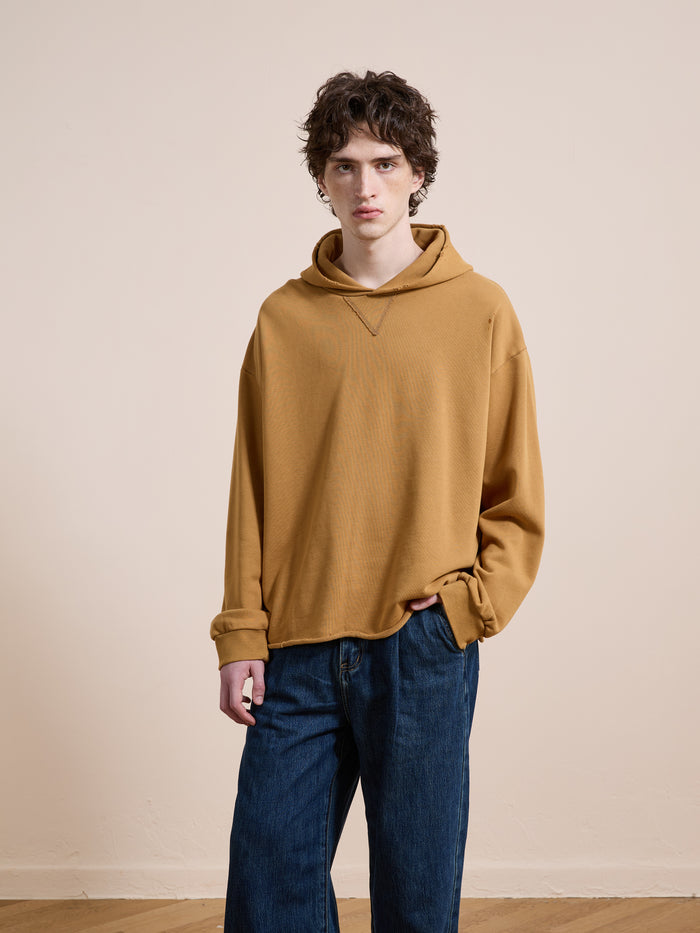 A person wearing the FOUND Beadwork Logo Hoodie in vintage enzyme wash mustard yellow and blue jeans stands against a plain beige wall, looking at the camera.