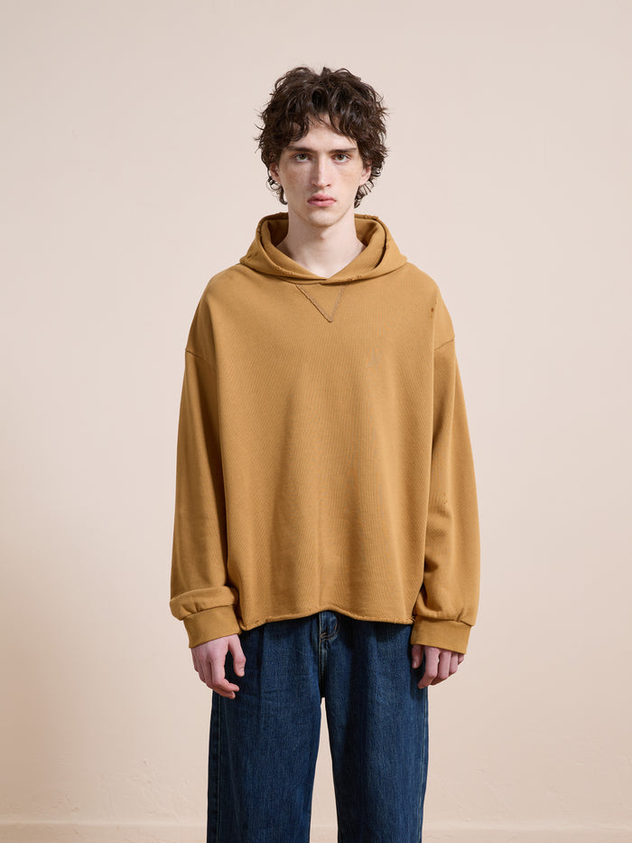 A person with curly brown hair wears the FOUND Beadwork Logo Hoodie in mustard yellow and blue jeans, standing against a plain beige background.