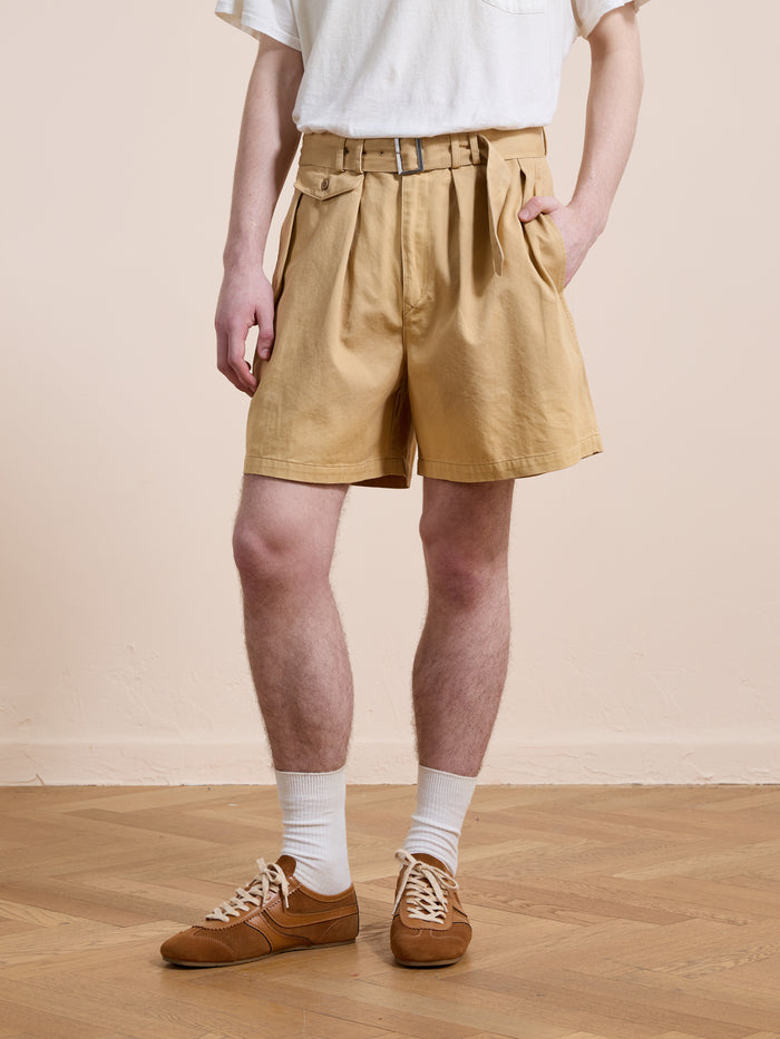 A person wearing a white t-shirt, brown Found Belted Twill Pleated Shorts, white socks, and brown sneakers stands on a wooden floor against a light beige wall.