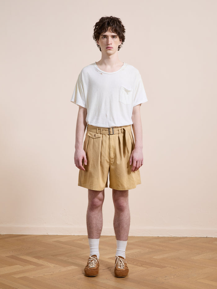 A person stands indoors against a plain wall, wearing a white t-shirt, Found Belted Twill Pleated Shorts in tan, white socks, and brown shoes.