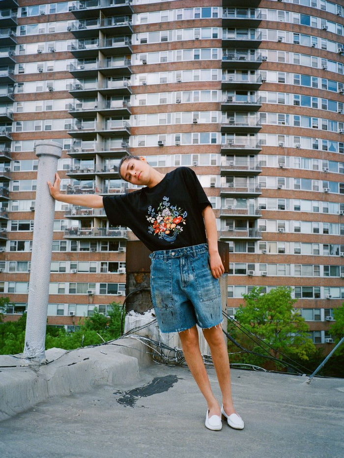 Wearing the FOUND Bouquet Flowers Tee and denim shorts, a person stands on a rooftop, leaning against a white pipe with a tall apartment building in the background.