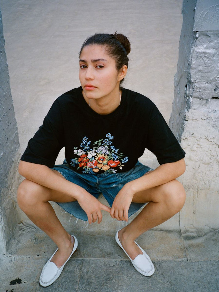 A person with dark hair in a bun is squatting outdoors, wearing the FOUND Bouquet Flowers Tee with Phulkari embroidery, denim shorts, and white slip-on shoes.