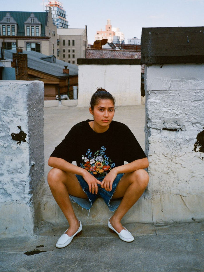 A person with hair in a bun squats on a rooftop, wearing the FOUND Bouquet Flowers Tee, denim shorts, and white slip-on shoes with urban buildings visible in the background.