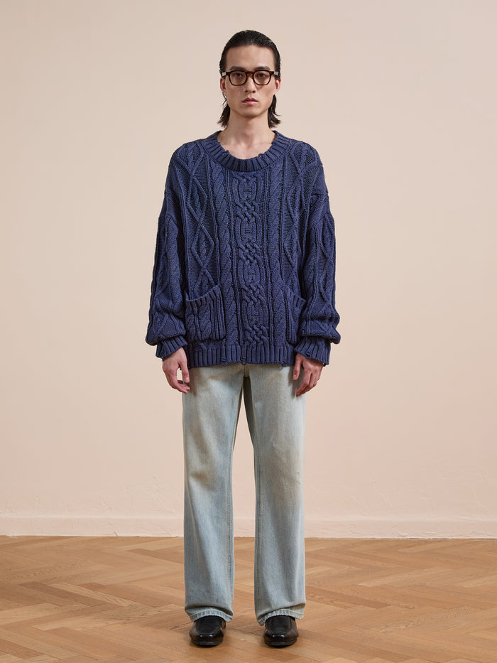 An individual wearing glasses, the Found Cable Knit Pullover Sweater in blue with pockets, paired with light-wash jeans and black shoes, stands indoors on a wooden floor against a plain background.