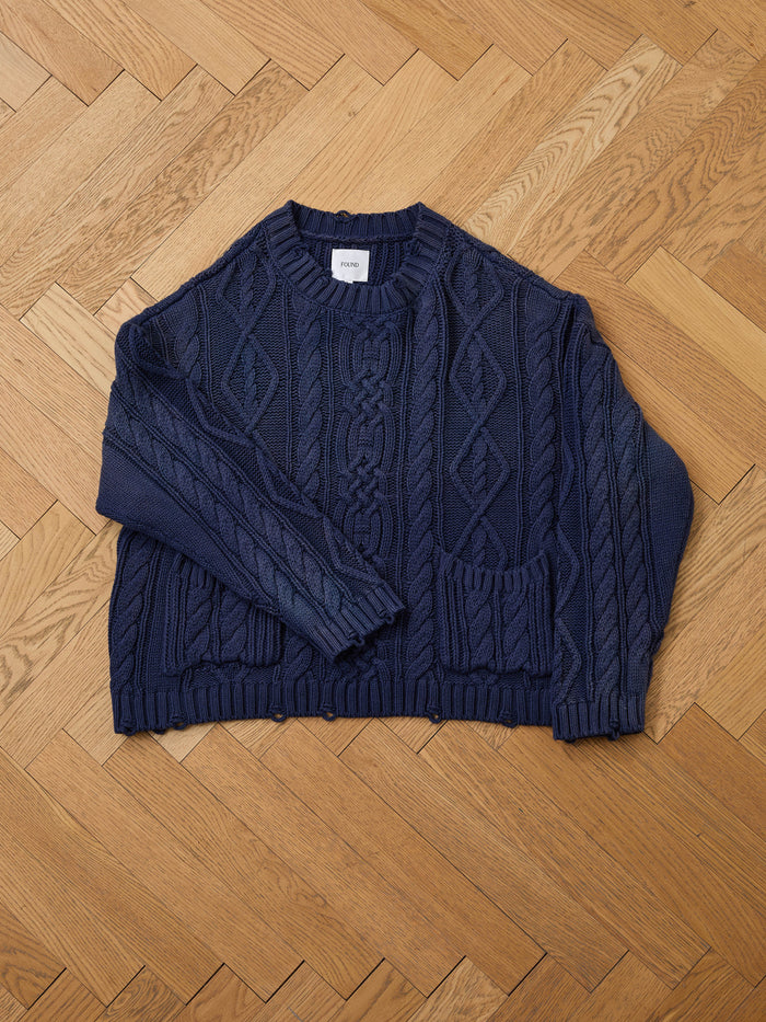 The Found Cable Knit Pullover Sweater, a unisex navy design with long sleeves and two front pockets, is laid flat on a herringbone wooden floor.