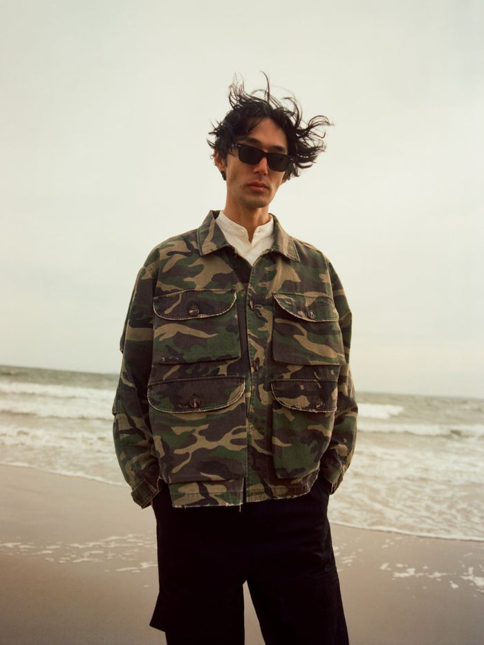 Wearing the FOUND Camouflage Twill Utility Cargo Jacket and sunglasses, a person stands on a beach as waves roll in under a cloudy sky—the midweight cotton twill brings rugged style to the coastal backdrop.