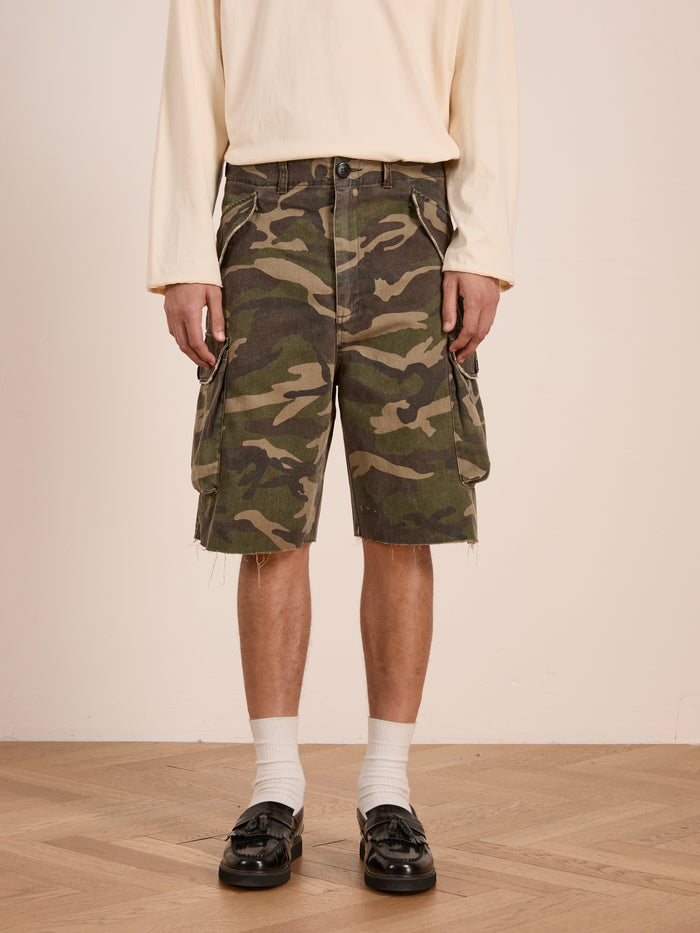 A person wearing a cream long-sleeve shirt, FOUND Camouflage Twill Utility Cargo Shorts, white socks, and black loafers stands on a wooden floor against a plain background.