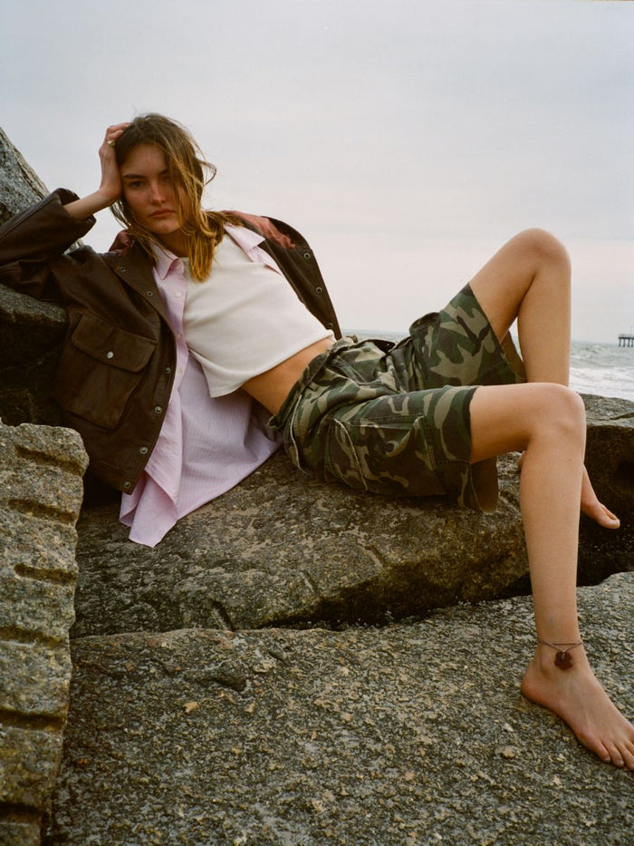 A woman sits barefoot on rocks by the sea, wearing a brown jacket, white crop top, pink shirt, and FOUND Camouflage Twill Utility Cargo Shorts for a vintage look.