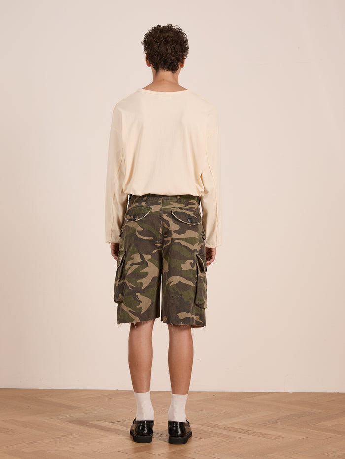 A person stands indoors facing away, wearing a beige long-sleeve shirt, FOUND Camouflage Twill Utility Cargo Shorts, white socks, and black shoes on a wooden floor.