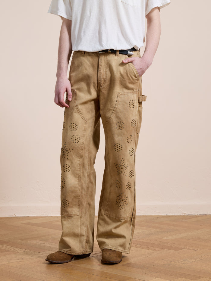 A person wears a plain white T-shirt, black belt, brown shoes, and FOUND Canvas Studded Work Pants while standing on a wooden floor against a beige wall. This unisex workwear look blends comfort with functional style.