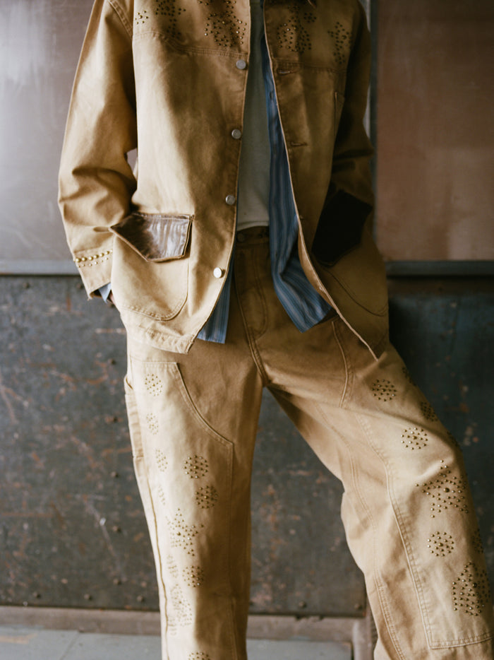 A person models the Found Canvas Studded Barn Jacket, a tan two-piece with laser-cut patterns and pockets, styled over a blue striped shirt and white top for a casual, workwear-inspired look. Face not visible.