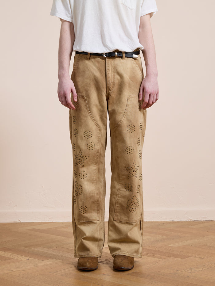 A person stands on a wooden floor against a plain light-colored wall, wearing FOUND Canvas Studded Work Pants with a patterned design, a white t-shirt, and brown shoes.
