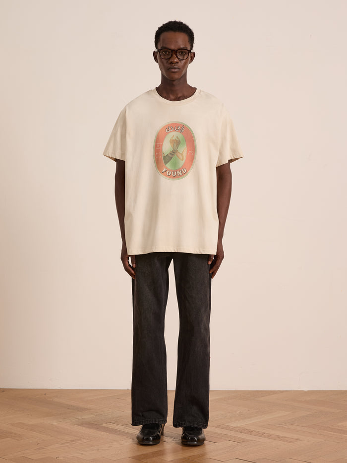 Indoors, someone wears glasses and the FOUND Community Hands Tee—a light-colored, oversized unisex cotton shirt with vintage matchbox artwork and "FOUND IN UNITY" text—paired with dark jeans and black shoes against a plain backdrop.