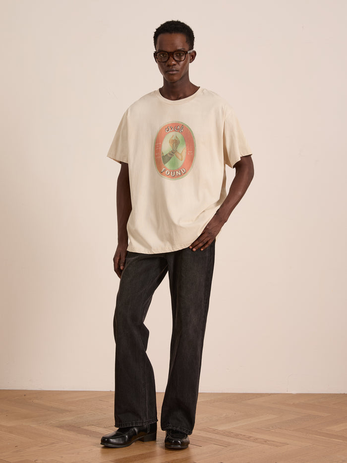 Indoors, a person models the FOUND Community Hands Tee—a cream unisex cotton shirt with vintage matchbox art—paired with dark jeans, black shoes, and glasses, against a plain light wall and wooden floor.