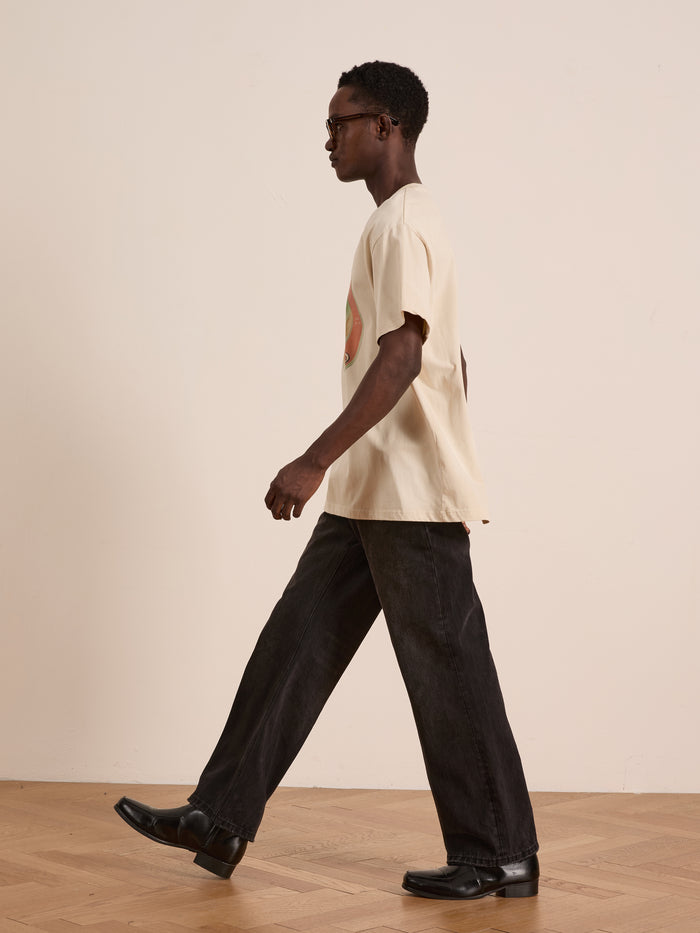 A man wearing the FOUND Community Hands Tee, black jeans, and black shoes walks indoors on a wooden floor against a plain light-colored wall.