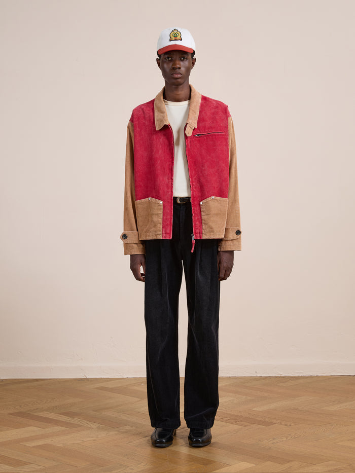 Indoors on a wooden floor, a person wears a white cap, Found’s Corduroy Field Jacket in red and tan panels, a white T-shirt, black trousers, and black shoes.