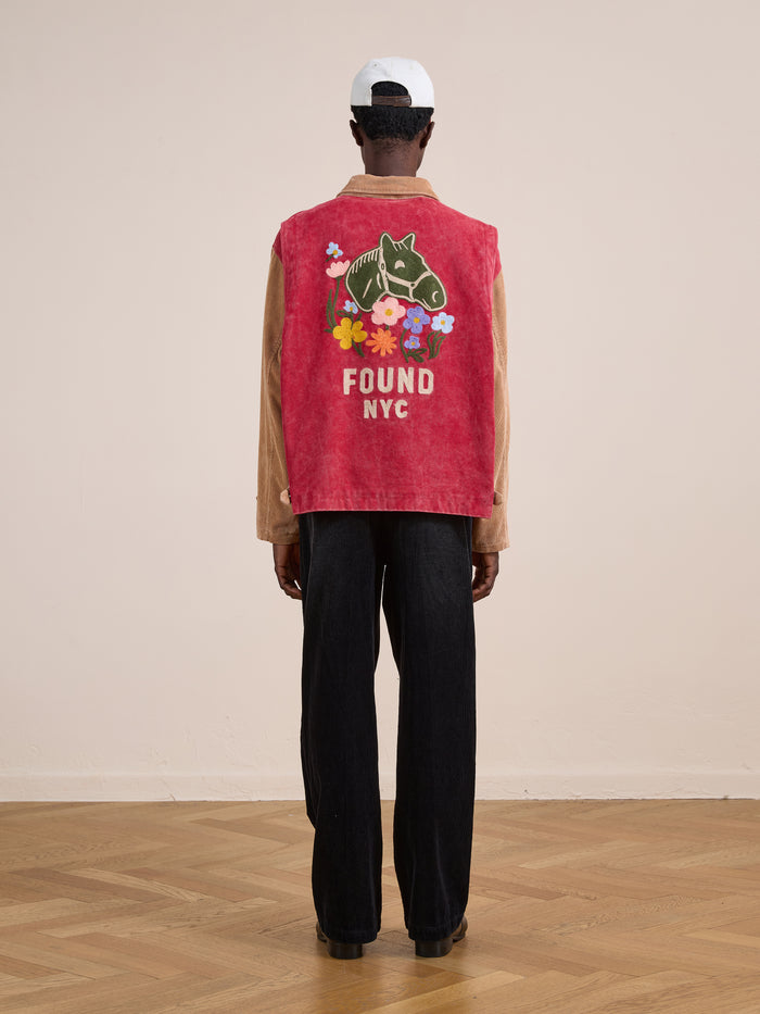 Person standing indoors, facing away, wearing the Found Corduroy Field Jacket in red with a horse graphic, colorful flowers, and "FOUND NYC" text on the back, paired with black pants and a white cap.