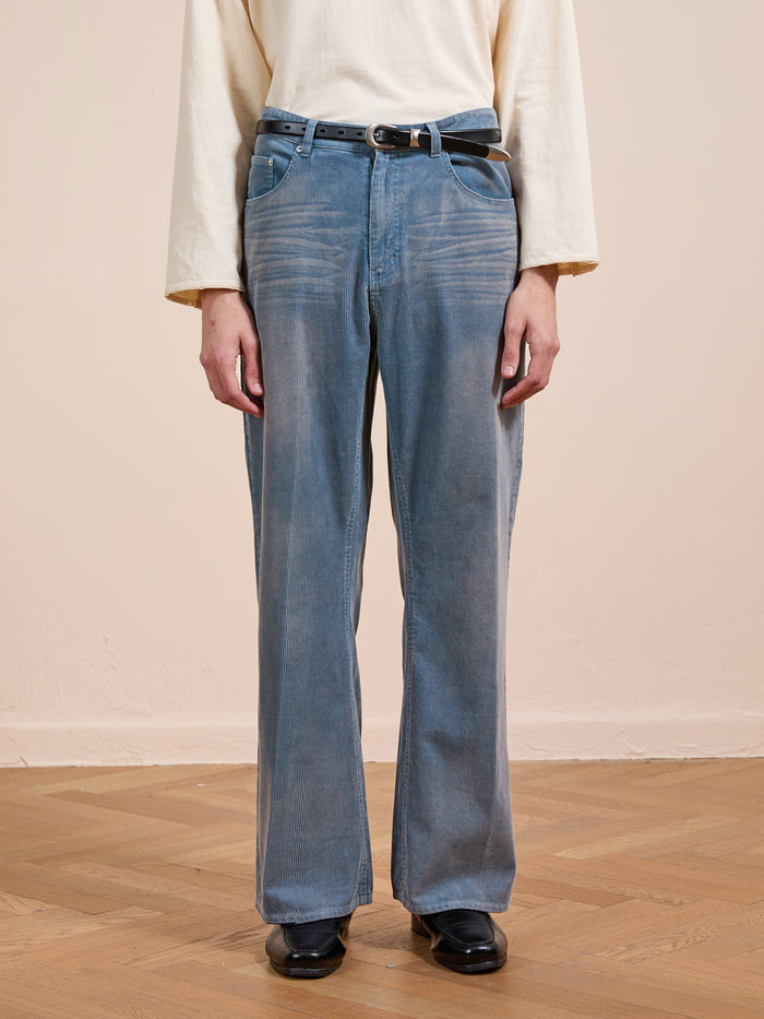 A person wears FOUND Corduroy Flare Work Pants in vintage blue, styled with a cream long-sleeve shirt, black belt, and black shoes, standing on a wooden floor against a beige wall.