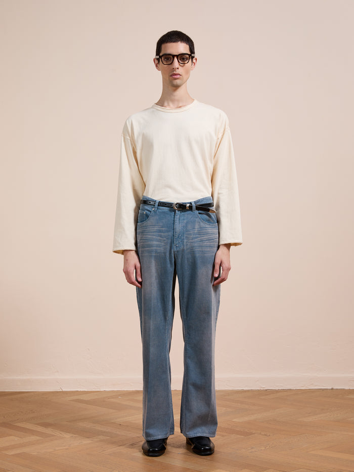 A person wears black glasses, an off-white long-sleeve shirt, FOUND Corduroy Flare Work Pants in vintage blue and a relaxed fit, and black shoes while standing on a wooden floor against a plain beige wall.