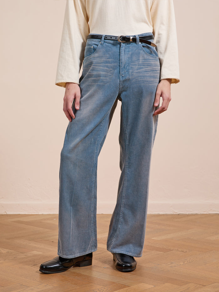 Person wearing FOUND Corduroy Flare Work Pants in vintage blue, paired with a cream long-sleeve shirt, black belt, and black shoes, standing on a wooden floor against a plain beige wall.