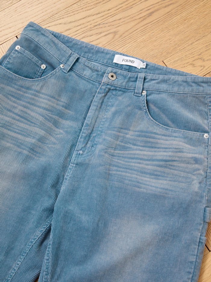 The FOUND Corduroy Flare Work Pants feature a vintage blue hue, front pockets, visible brand label, and a relaxed fit style, shown laid flat on a wooden floor.