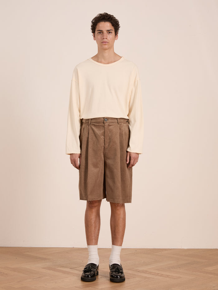 A person stands indoors against a plain background, wearing a cream long-sleeve shirt, FOUND Corduroy Pleated Trouser Shorts in brown (unisex relaxed fit), white socks, and black shoes.