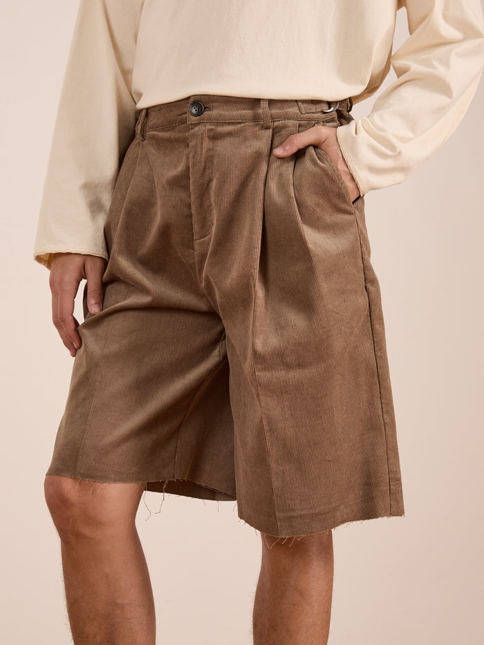 A person wearing a cream long-sleeve shirt and FOUND Corduroy Pleated Trouser Shorts in a relaxed unisex fit with double pleats and raw hems, standing with one hand in their pocket.