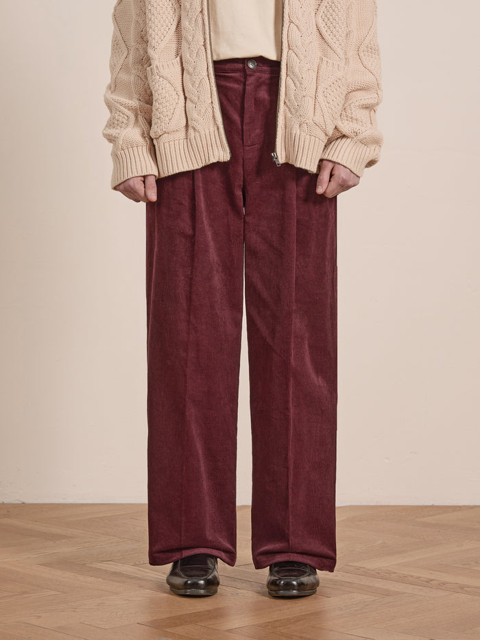 A person wearing a cream cable-knit cardigan, cream shirt, FOUND Corduroy Pleated Trousers in a burgundy shade with a wide-leg silhouette and high-rise waist, and black shoes stands on a wooden floor.