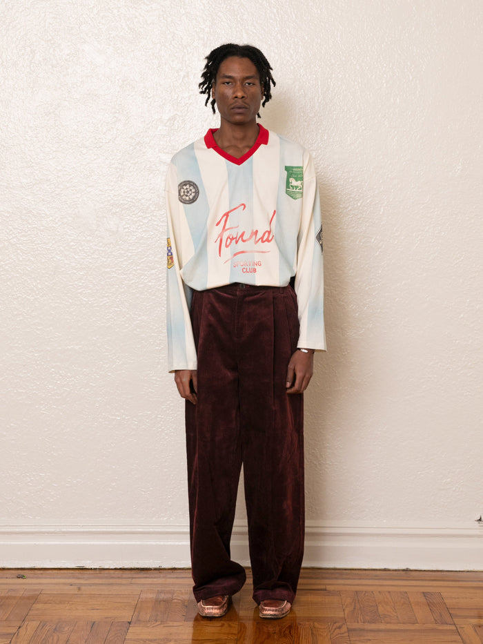A person stands indoors against a plain white wall and wooden floor, wearing a white long-sleeve shirt with red lettering paired with FOUND Corduroy Pleated Trousers. The trousers feature a wide-leg silhouette, dark brown corduroy fabric, and a high-rise waist.