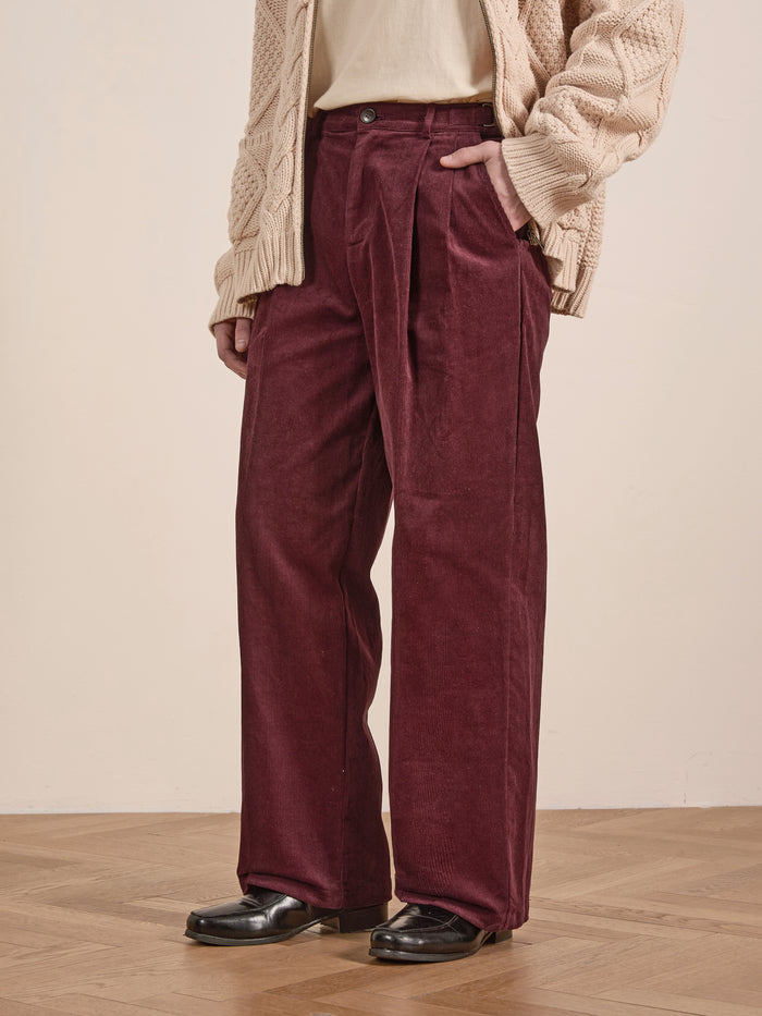 A person wearing FOUND Corduroy Pleated Trousers in maroon with a high-rise waist and wide-leg silhouette, paired with a cream sweater and black shoes, stands on a wooden floor with one hand in their pocket.