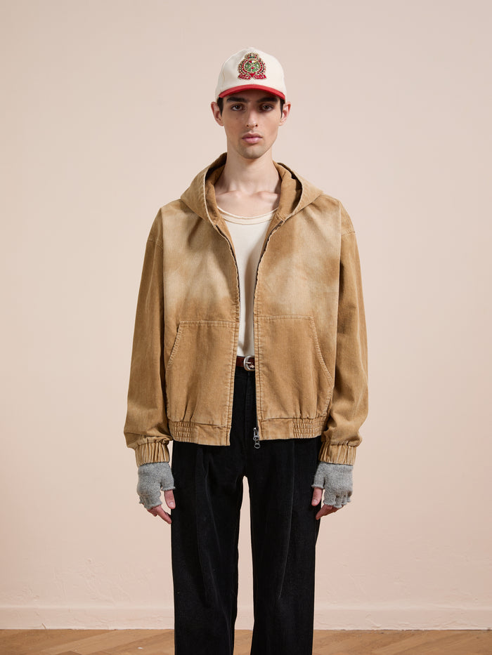 A person stands against a plain background wearing the FOUND Corduroy Zip Up Hoodie in beige, styled with a white shirt, black pants, fingerless gloves, and a white baseball cap featuring a red brim and embroidered patch for a vintage look.